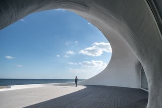The UCCA Dune Art Museum looks out to the sea. The structure is open and curved with wooden decking. A man is standing on the deck and is looking out to the sea.