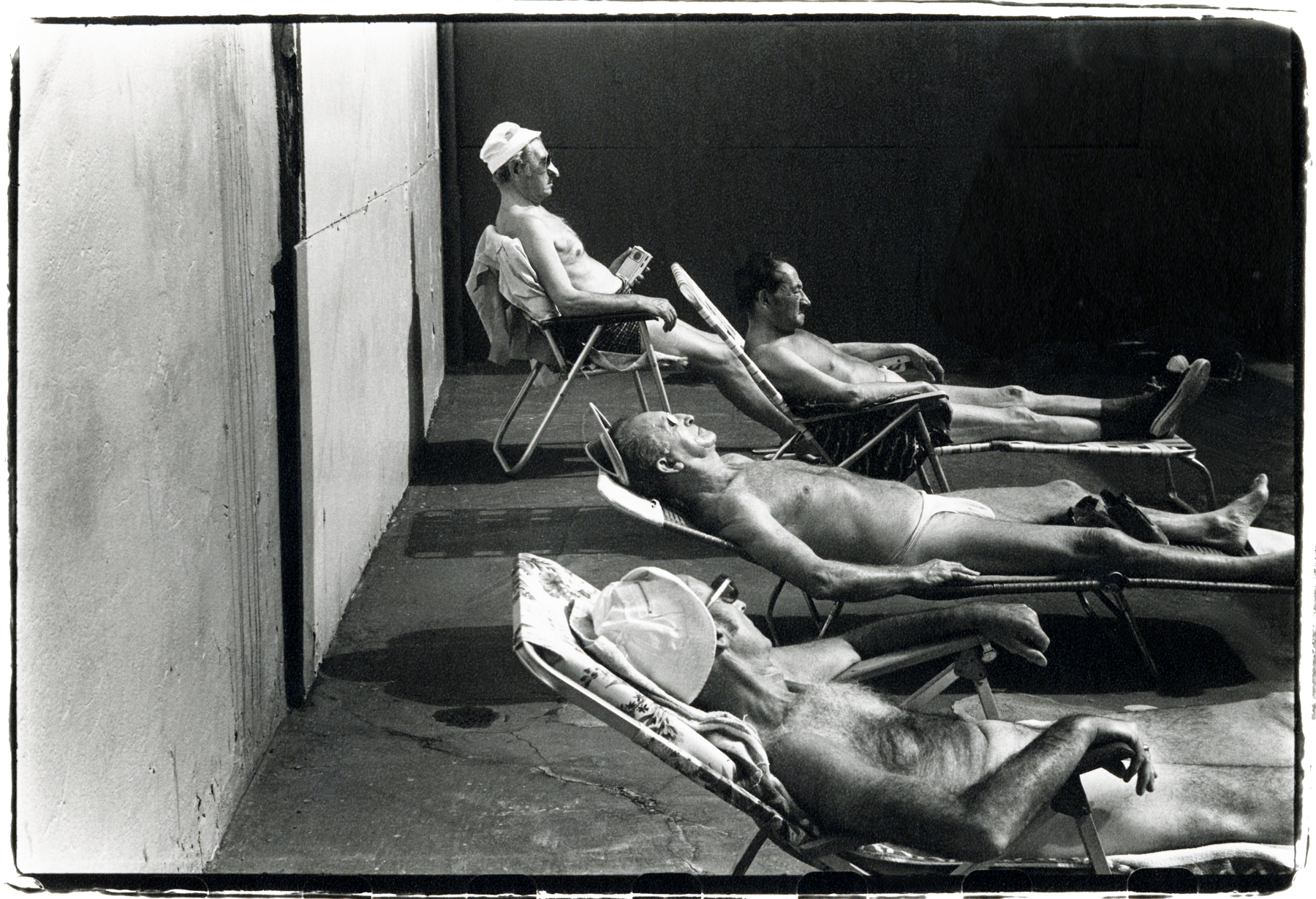 Four older men sunbathe in deckchairs and loungers against a stark wall, their faces turned up to the light.