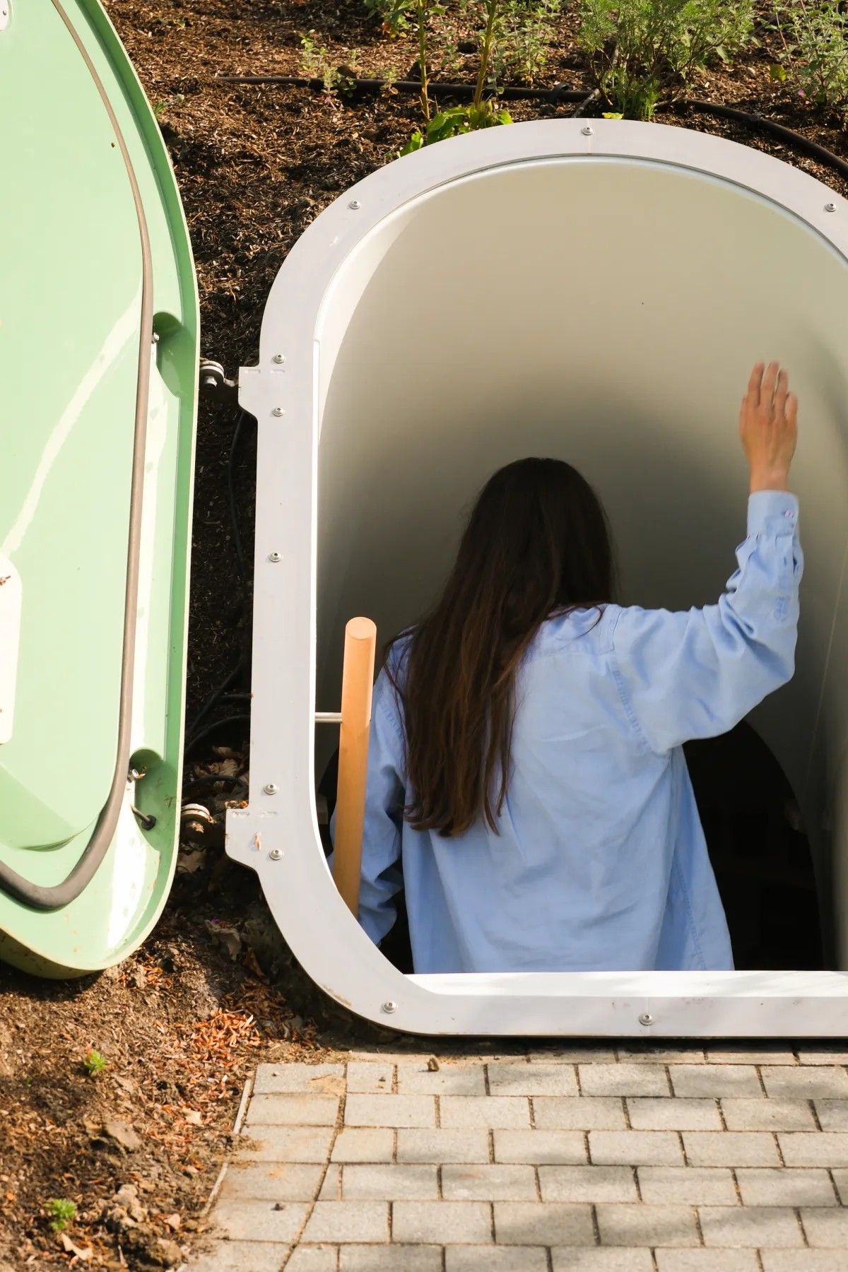 A young woman in a blue shirt and denim jeans opens the lacquered green door to a subterranean fridge in a garden, where a series of pickle jars sits.