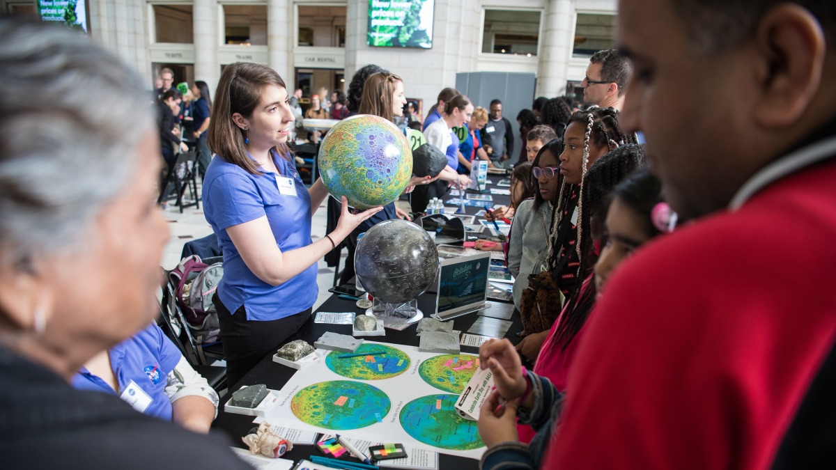 people gather around a series of exhibits. A person in the center is holding a globe up to the audience.