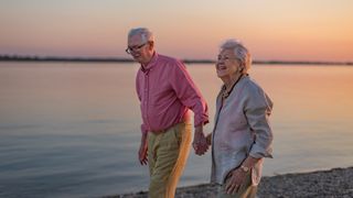 A senior man and woman smile as they walk along the beach hand in hand during a sunset