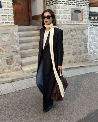 Danish influencer Ilirida Krasniqi walking on a sidewalk wearing black sunglasses, a long beige scarf, long maxi black coat, white T-shirt, dark wide-leg jeans, and black boots