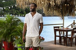 Emmanuel Warner (Bobby Gordon) stands at Catherine's Bar in the outside seating area, with the ocean and some lush green trees visible behind him. He is wearing a translucent white linen shirt and a pair of khaki shorts. He is smiling and has his left hand in his pocket.