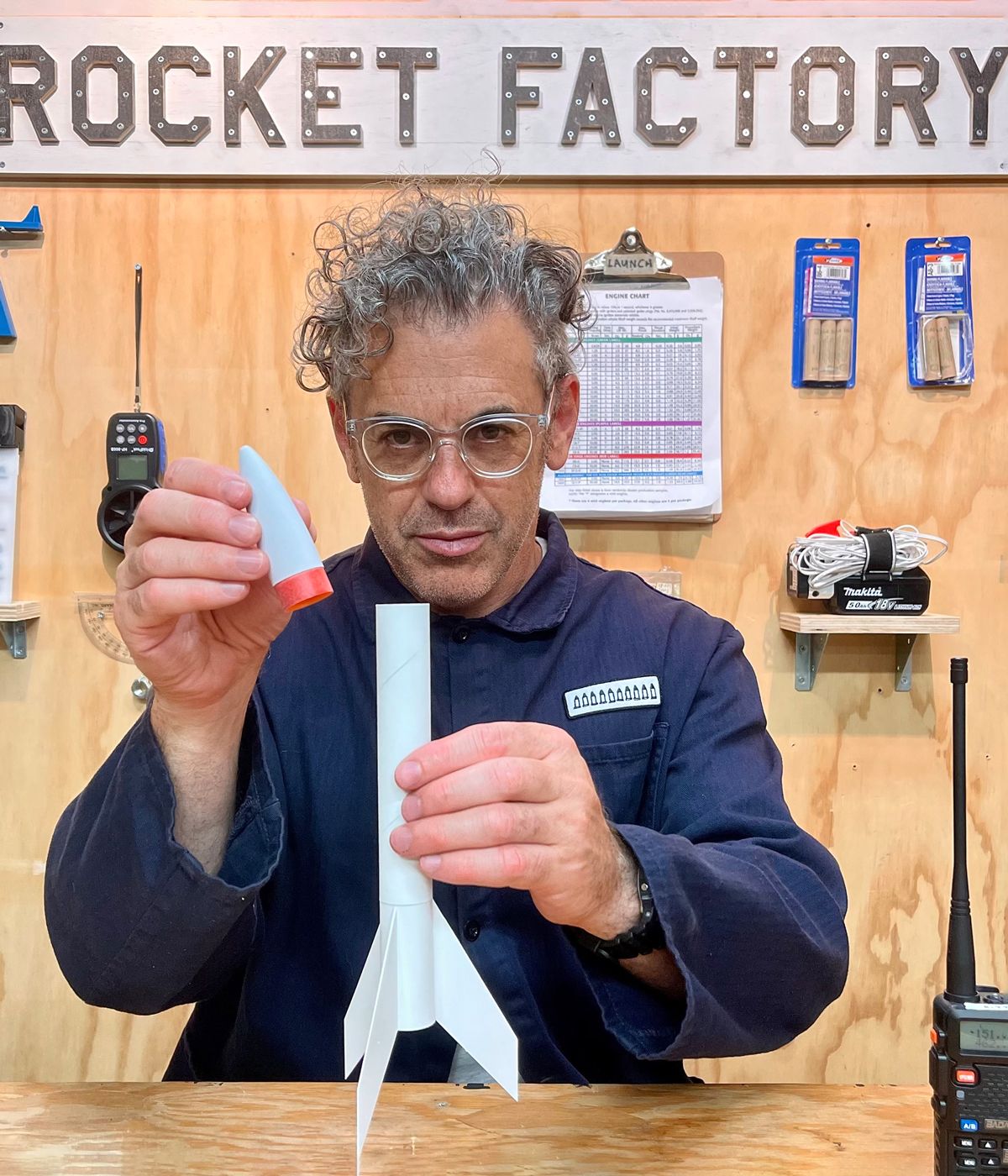 Tom Sachs building a white model rocket, wooden worktop surface, remote control device, wooden oanel behind him with small wooden shelves with display items