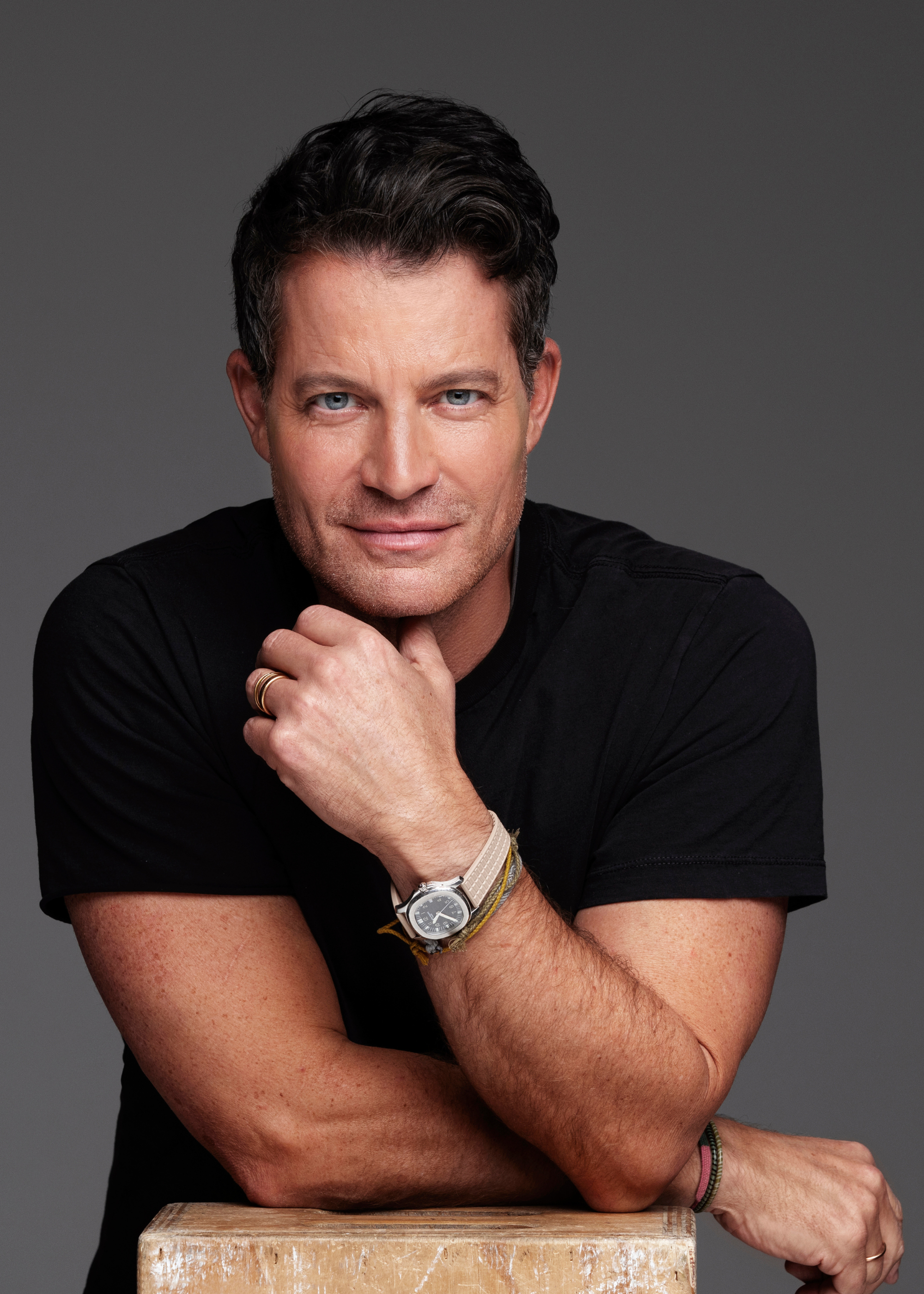 Nate Berkus headshot leaning on a plinth with one arm resting on the plinth and the other touching his chin while smiling at the camera wearing a black t-shirt
