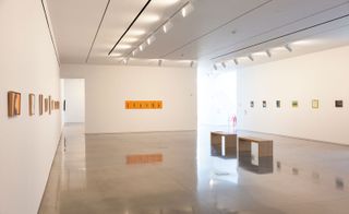 A gallery with small paintings on white walls and seating benches in the middle of a shiny floor.