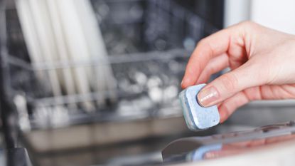 A hand holding a dishwasher tablet in front of an open dishwasher