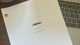 Photo of the finished script of Babylon 5's new movie. Here we see a white page which reads "Babylon 5:" with the later half of the title scribbled out. Underneath this is black text "Written by J. Michael Straczynski. The script has been played on top of a laptop keyboard on a dark wooden desk.