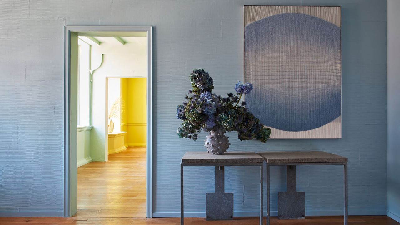 light blue wall with doorway leading into light green room, gray metal tables with vase of flowers and abstract blue artwork on the wall