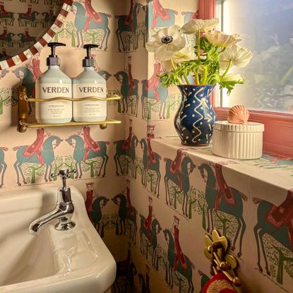 Corner of bathroom with horse patterned wallpaper, hand wash and lotion in gold brass holder and small vase of flowers on window sill