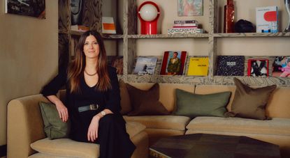 A woman with long straight brown hair wears black trousers, a matching top, and a large belt while sitting atop a velvety beige couch with green, brown pillows and books displayed behind it.
