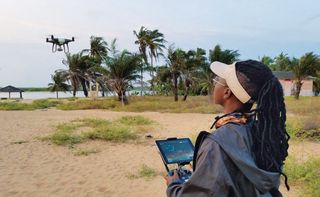 Woman stood in a sandy area controlling an airborne drone, palm trees swaying, water and small building in the distance, cloudy sky