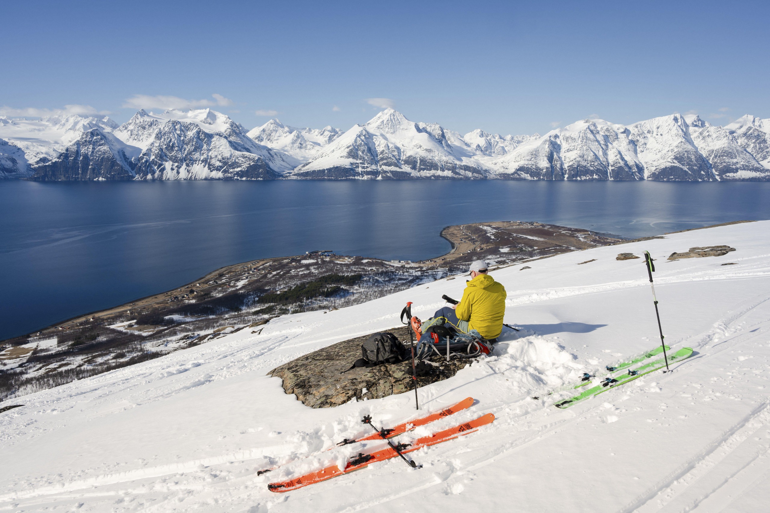 A ski tourer sits on a chair, using a rock as a desk, while looking out over a fjord