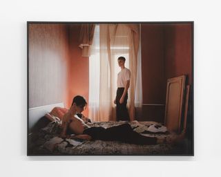 Two young people lying on a bed in a darkly lit room as another young man stands next to the bed watching them