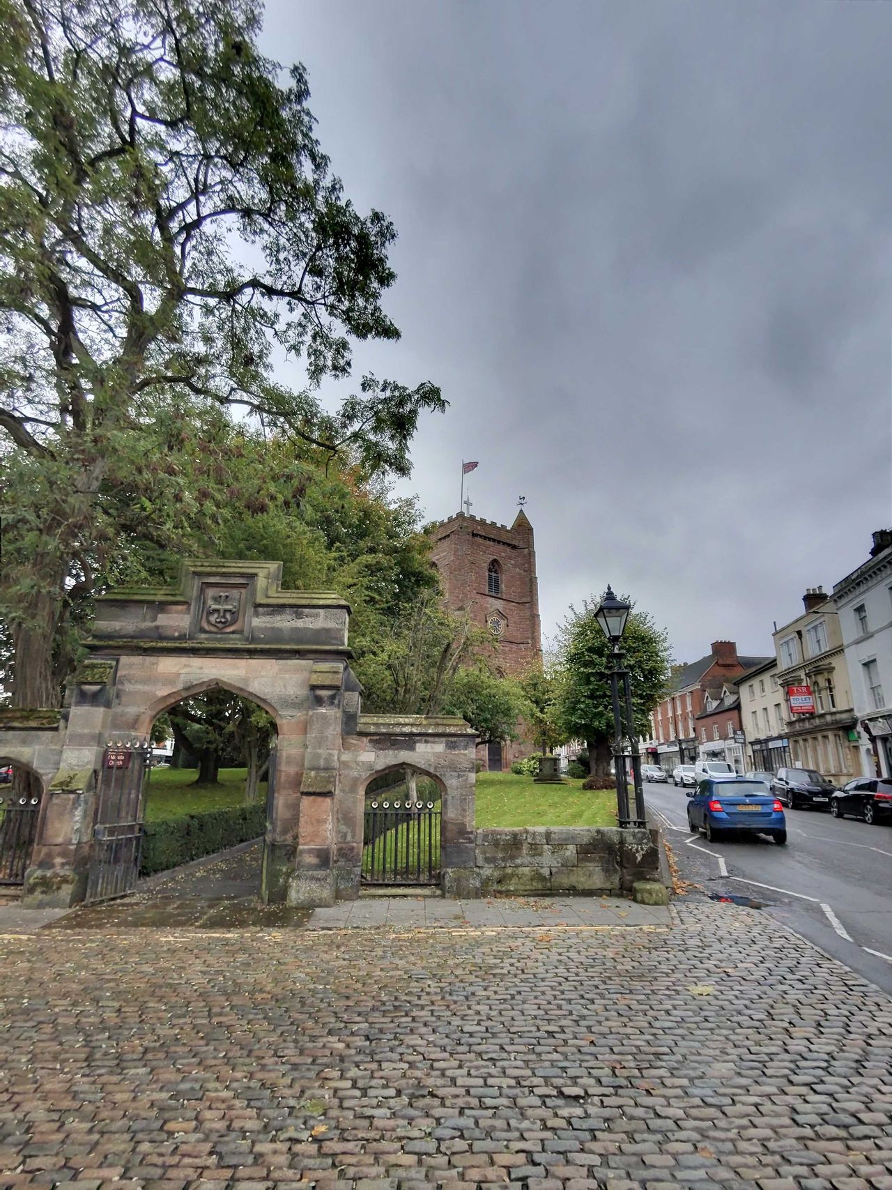 A church on a highstreet on a cloudy day, a flag flies above it and cars are driving past