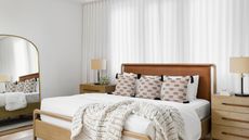 Bright bedroom with large wooden framed bed, dressed with white sheets and playful cushion covers, framed by white venetian blinds