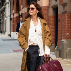Katie Holmes wears a trench coat, white button-up, black trousers, black square-toe loafers, and a burgundy alo bag.
