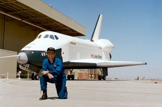 a man in a blue flight suit kneels in front of a white space shuttle on a runway
