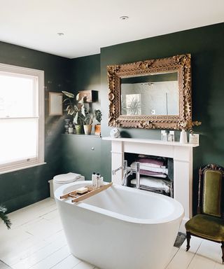 A white bath in the center of a bathroom with dark green walls and an ornate wall mirror