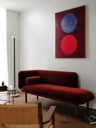 living room corner with modern floor lamp, artwork with purple and red circle, red velvet bench seat, and woven chair