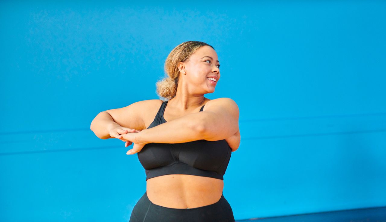 A woman in black sportswear rotates her upper body and looks over her shoulder. She stands in front of a bright blue wall