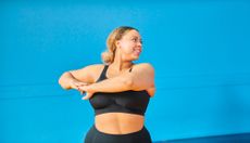 A woman in black sportswear rotates her upper body and looks over her shoulder. She stands in front of a bright blue wall