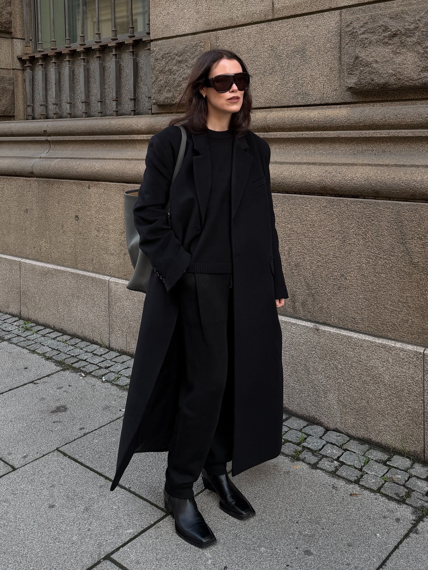 Annabel wears a black coat, black sweater, black trousers, black boots.