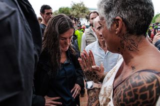 Queen Letizia looking down in sadness wearing a jacket covered in mud as she speaks to a woman covered in tattoos