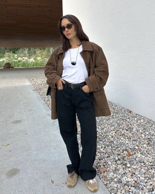 Fashion influencer Debora Rosa wearing black oval sunglasses, a black pendant necklace, white T-shirt, brown suede jacket, black belt, baggy black jeans, and Miu Miu brown suede sneakers.