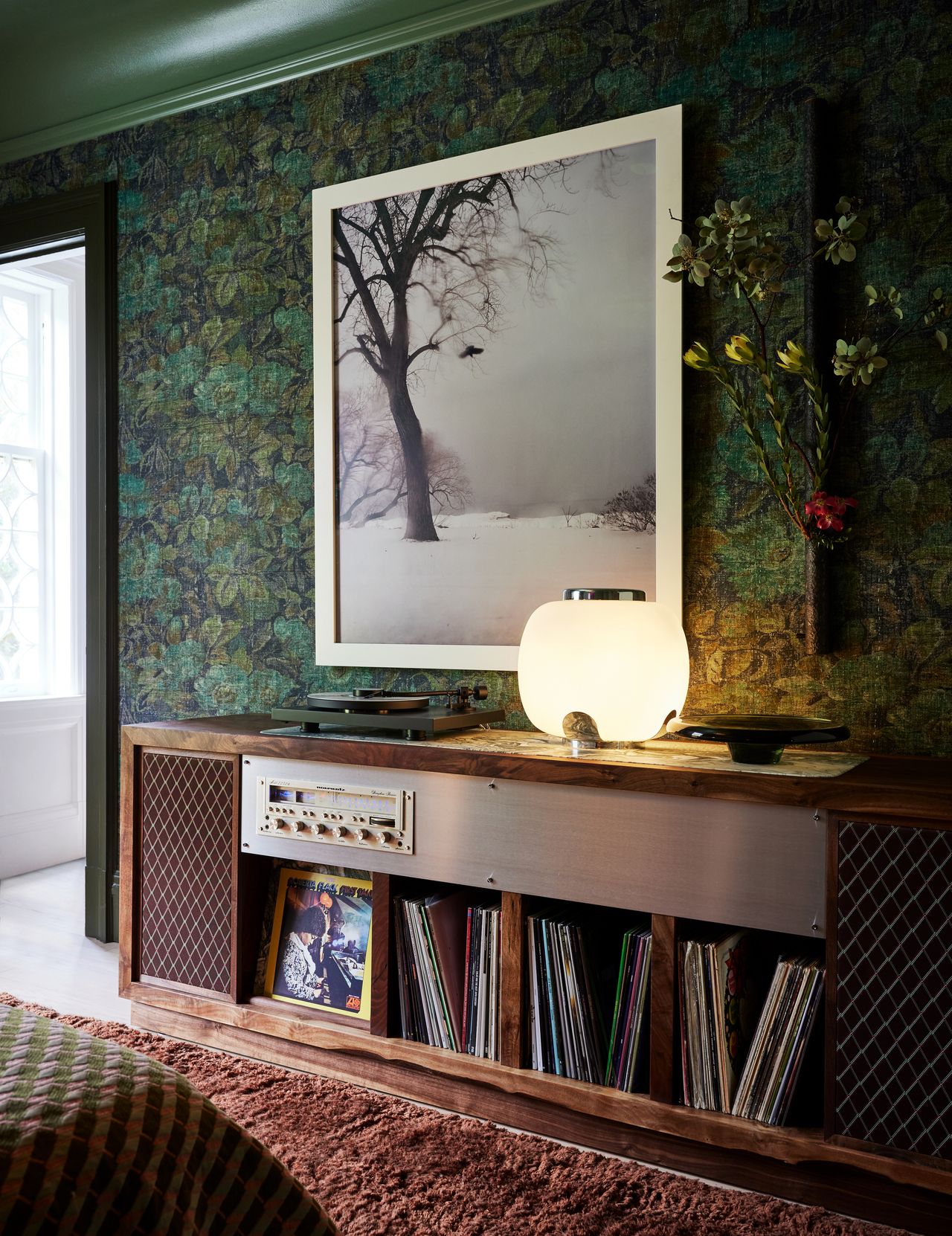 Image of a forest green bedroom that has a patterned wallpaper. The floor has a shaggy, burnt orange rug and a long and low wooden record storage cabinet and built-in speaker.