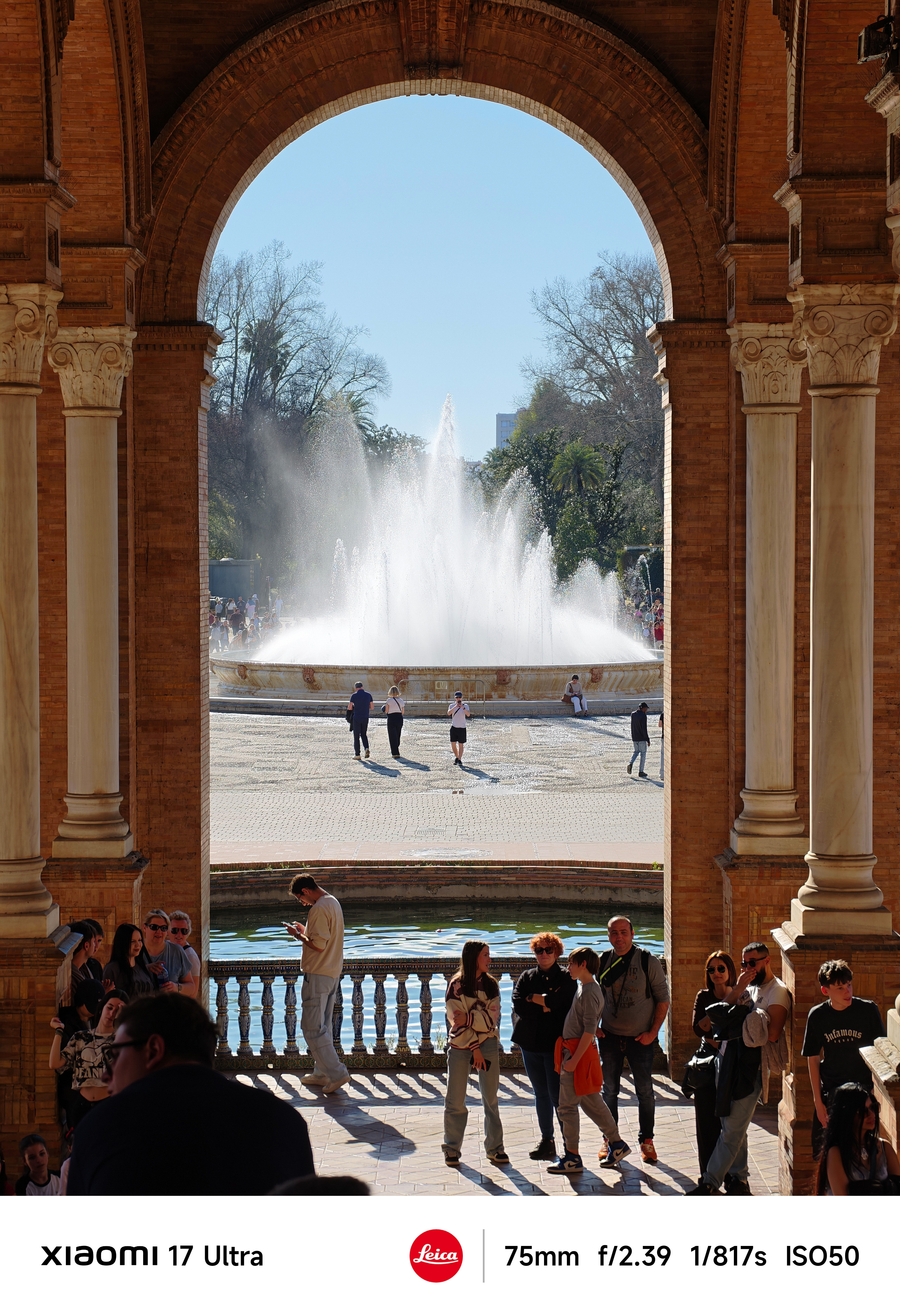 People gathered beneath a grand brick arch framing a large fountain in bright sunlight.