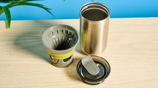 Wacaco Cuppamoka, a stainless steel coffee pour-over brewer, photographed against a blue background, with brown filters and a sippy lid