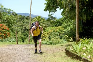 Jonny (Simon Lennon) is wearing workout gear and a heavy-looking backpack, making his way up a mud trail surrounded by luscious greenery. He's covered in dirt, and is limping on his right leg.