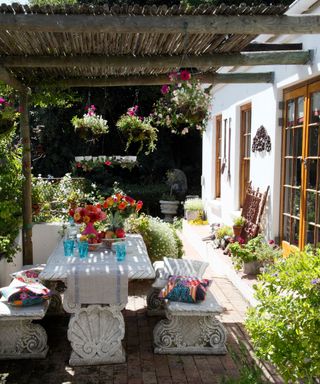 Ornate stone patio furniture under a rustic wooden pergola, with colorful cushions and cut flowers.