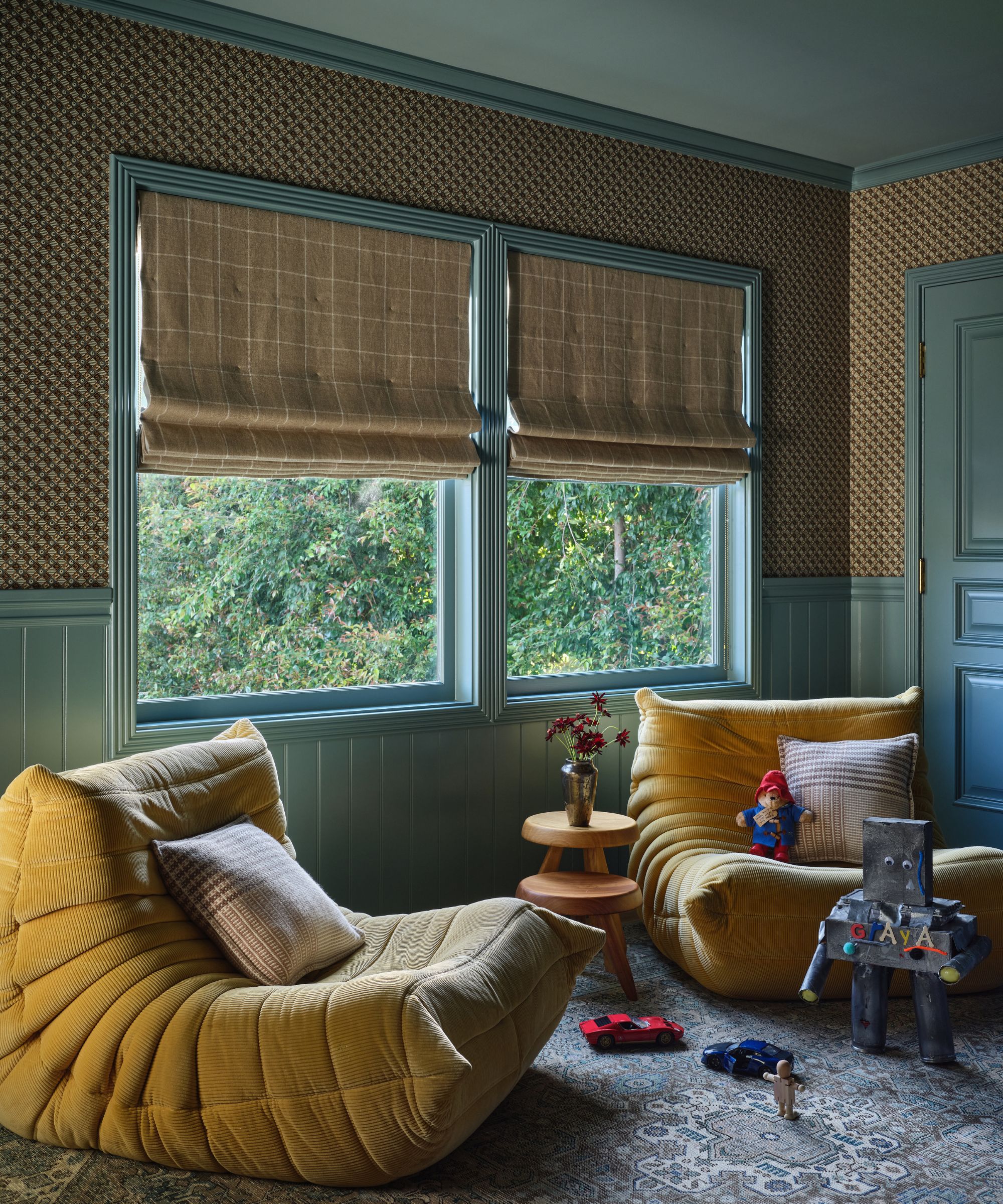 Children&#039;s sitting area with olive green walls and wainscoting, two large, slouchy yellow corduroy bean bag chairs, and windows with checkered Roman blinds