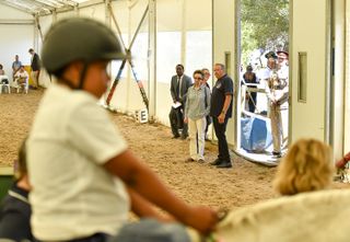 Princess Anne wearing white pants and a blue top standing next to two men watching a child riding a horse in a dirt ring