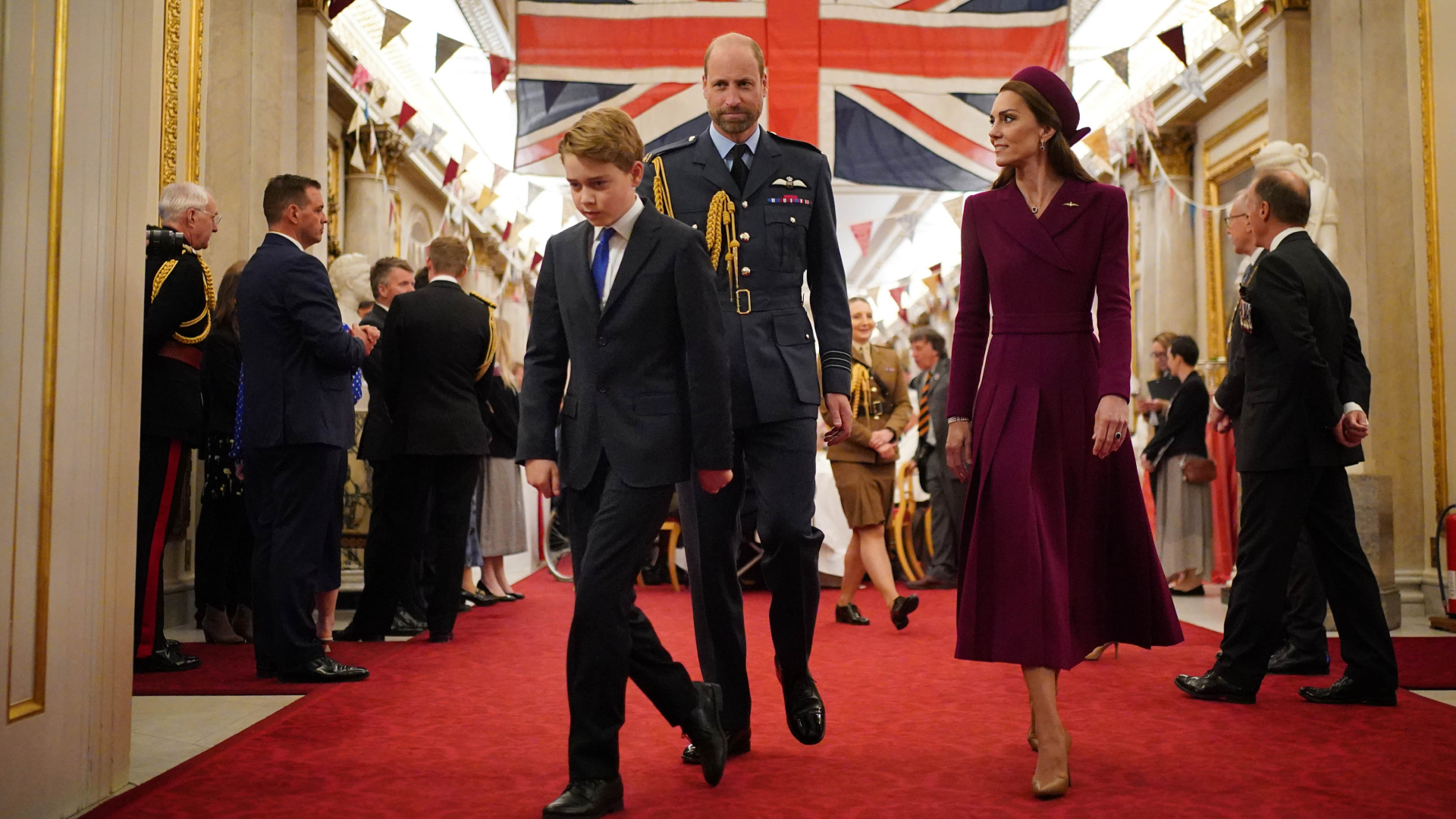 Prince George walking in front of Prince William in a military uniform and Prince Kate in a burgundy coat with a Union Jack behind them