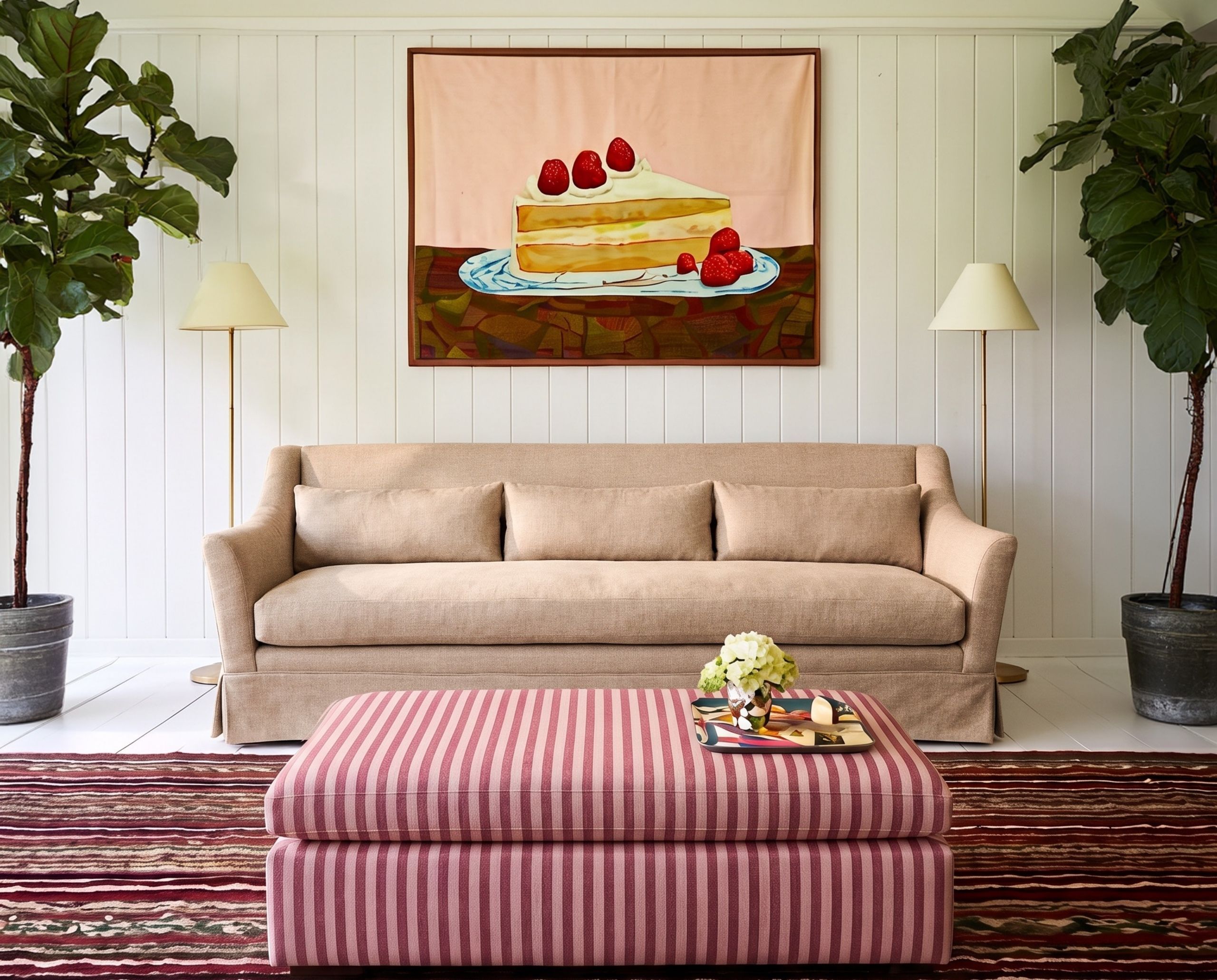 A tan three-seater sofa in a white living room with a painting of a cake above it and a pink striped ottoman in front of it.