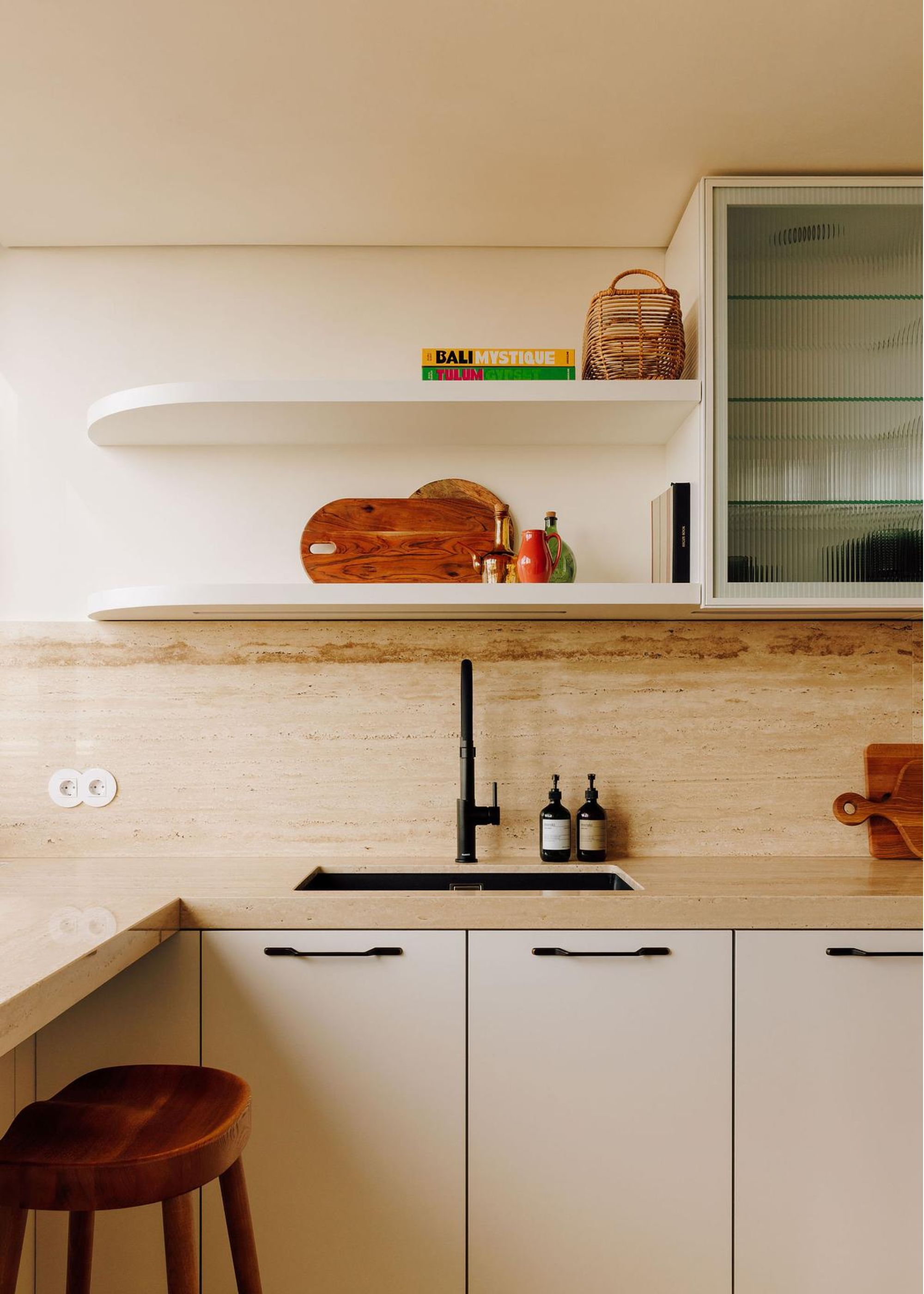 The corner of a kitchen with a cream colored countertop that lead to the splashback. There is also a sink with a tap as well as handwash and dish soap beside it. Above the are two open shelves with kitchen accessories on top