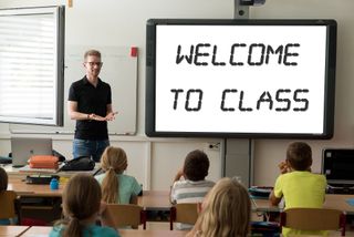 A teacher stands in front of a classroom next to a screen with the words "Welcome to Class" on it. 