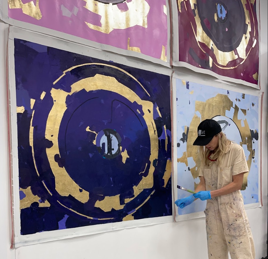 Kobieta w kombinezonie stoi obok czterech kolorowych obrazów przedstawiających właz statku kosmicznego