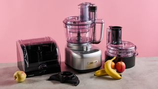 The Cuisinart Expert Prep Pro food processor pictured against a pink background. The accessories storage case is pictured to its left, and the dicing accessory kit is pictured on its right. The stone-effect tabletop that it sits on has apples and bananas on.