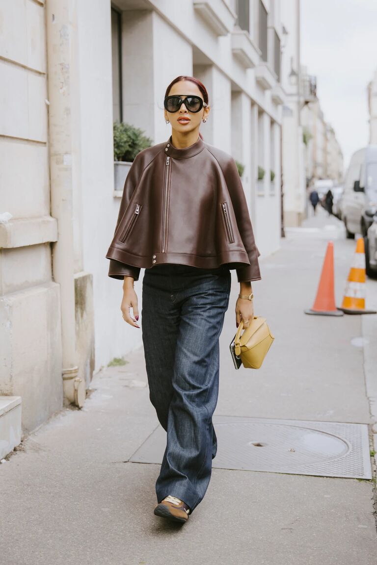a woman at fashion week wearing a brown leather jacket, baggy jeans, and brown suede sneaker