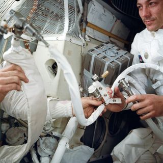 After an explosion damaged the service module, the Apollo 13 astronauts had to use the lunar module as a "lifeboat" to get home. Systems in the lunar module had to be rigged to make it possible to get the men back to Earth. Hoses were connected to various apparatus to make the trip home possible. The astronauts pictured here are holding the feed water bag from the portable life support system, which is connected to a hose from the Lunar Topographic Camera. Behind the astronauts the "mail box" can be seen — a creation built by the crewmen using the command module's lithium-hydroxide canisters that clean carbon dioxide from the air inside the spacecraft.
