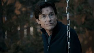 Jason Bateman sitting on a swing, looking to his left, towards the camera. 