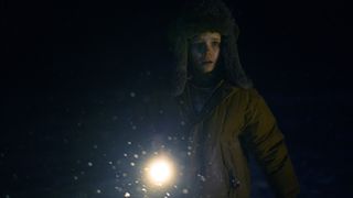 A young girl holding a flashlight, searching in the dark. She is wearing a thick winter hat and jacket.