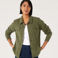 Middle-aged woman wearing green jacket, white top and blue denim jeans, with white backdrop behind her.