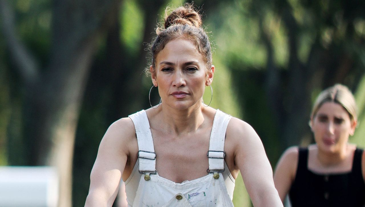 Jennifer Lopez bikes with a friend in the Hamptons while wearing paint splattered overalls, a high bun, and large hoop earrings