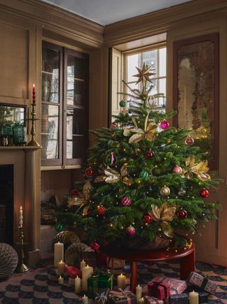 Christmas tree with gold, pink, green, and red decorations in a gold/beige living room corner near a fireplace. There are presents and candles under the tree.