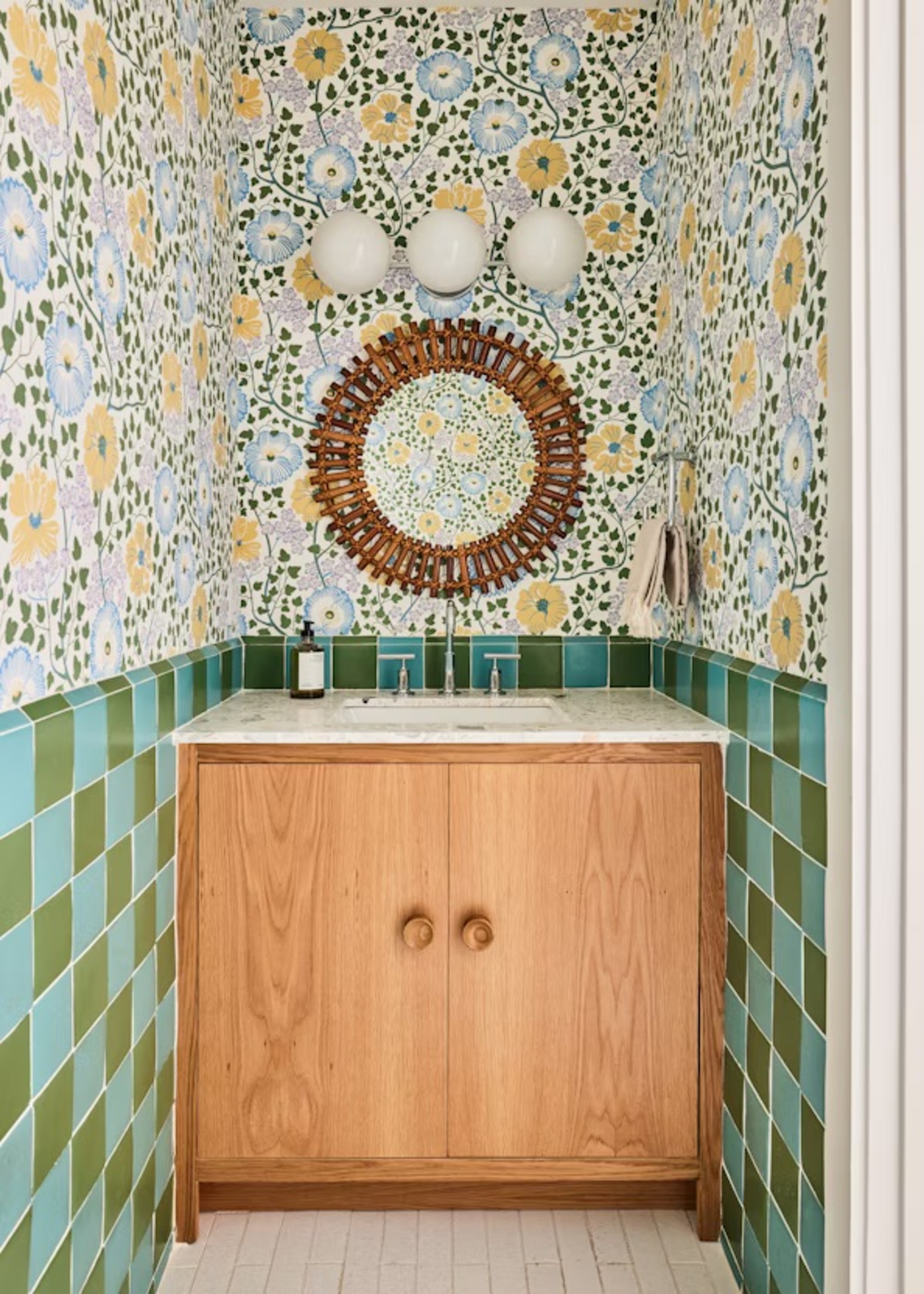 colorful bathroom with green checkered tiles and colorful floral wallpaper with a bamboo round mirror, three wall sconces, timber built-in vanity with marble counter and hand soap